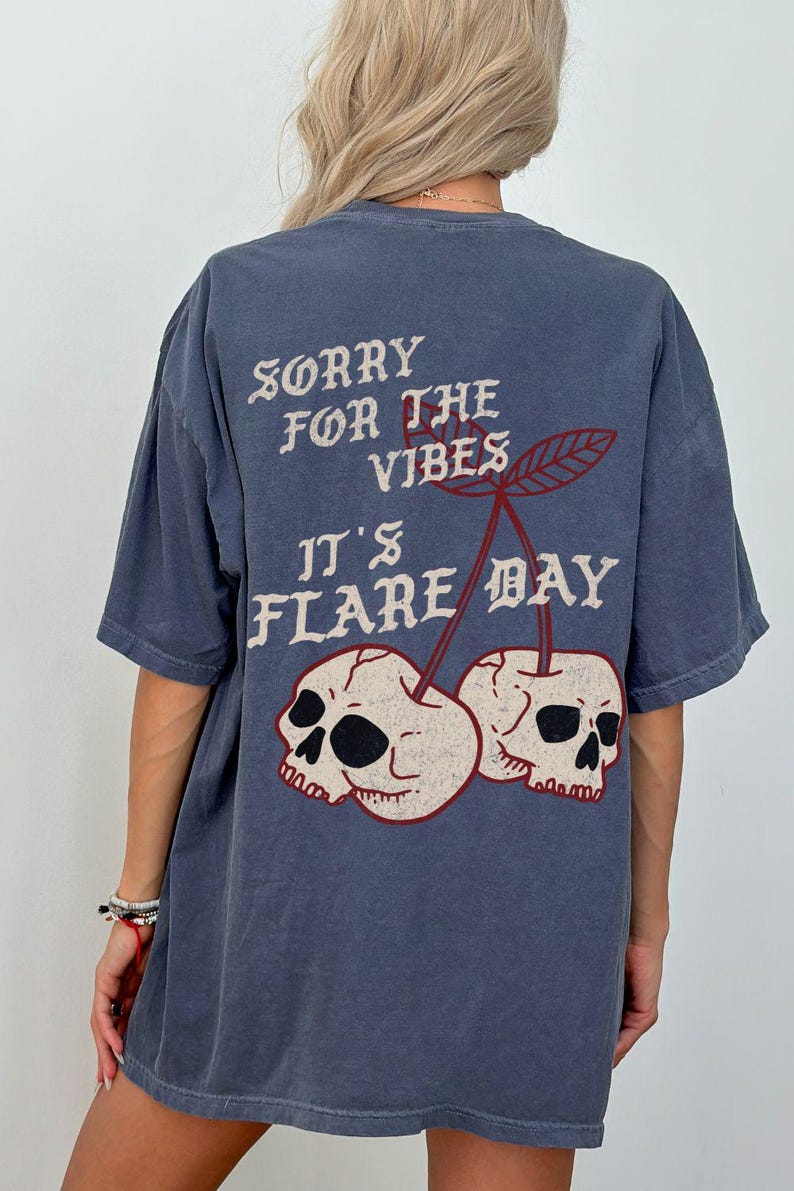 May include: A blue t-shirt with the text "SORRY FOR THE VIBES IT'S FLARE DAY" in white and red. The design features two skulls in the shape of cherries. The shirt is a casual, oversized fit.