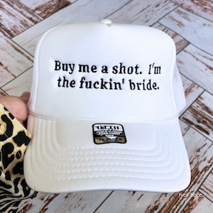 May include: White trucker hat with the embroidered text "Buy me a shot. I'm the fuckin' bride." The hat has a white foam front and a white mesh back. A small black and white tag is visible on the front.