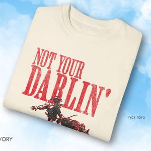 Puede incluir: Una camiseta blanca con el texto "NOT YOUR DARLIN'" en letras rojas. Debajo del texto se ve un vaquero montando un caballo.