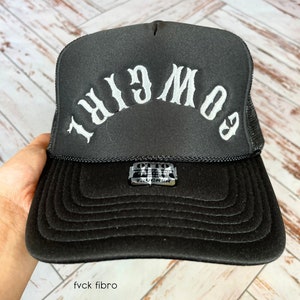 May include: Black trucker hat with a white embroidered logo that reads "GOOD GIRL" in a backwards font.