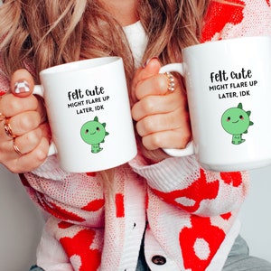 Pu&ograve; includere: Due tazze in ceramica bianca con un motivo di dinosauro verde e il testo "felt cute might flare up later, idk".
