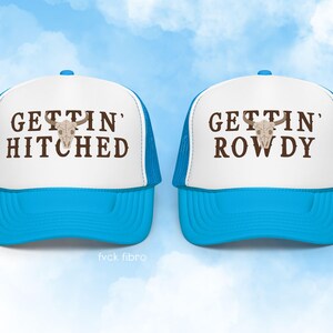 K&ouml;nnte beinhalten: Zwei wei&szlig;e Trucker-Caps mit blauen Netzr&uuml;cken. Eine M&uuml;tze sagt "Gettin' Hitched" und die andere "Gettin' Rowdy". Beide M&uuml;tzen zeigen einen braunen Sch&auml;del mit H&ouml;rnern.