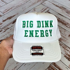 以下が含まれることがあります： 緑色の刺繍で「BIG DINK ENERGY」と書かれた白いトラッカーハット。帽子には、白い文字で「OTTO WORLD FAMOUS FOAM FRONT OSFM TRUCKER DURABLE SNAP CLOSURE」と書かれた黒いラベルが付いています。