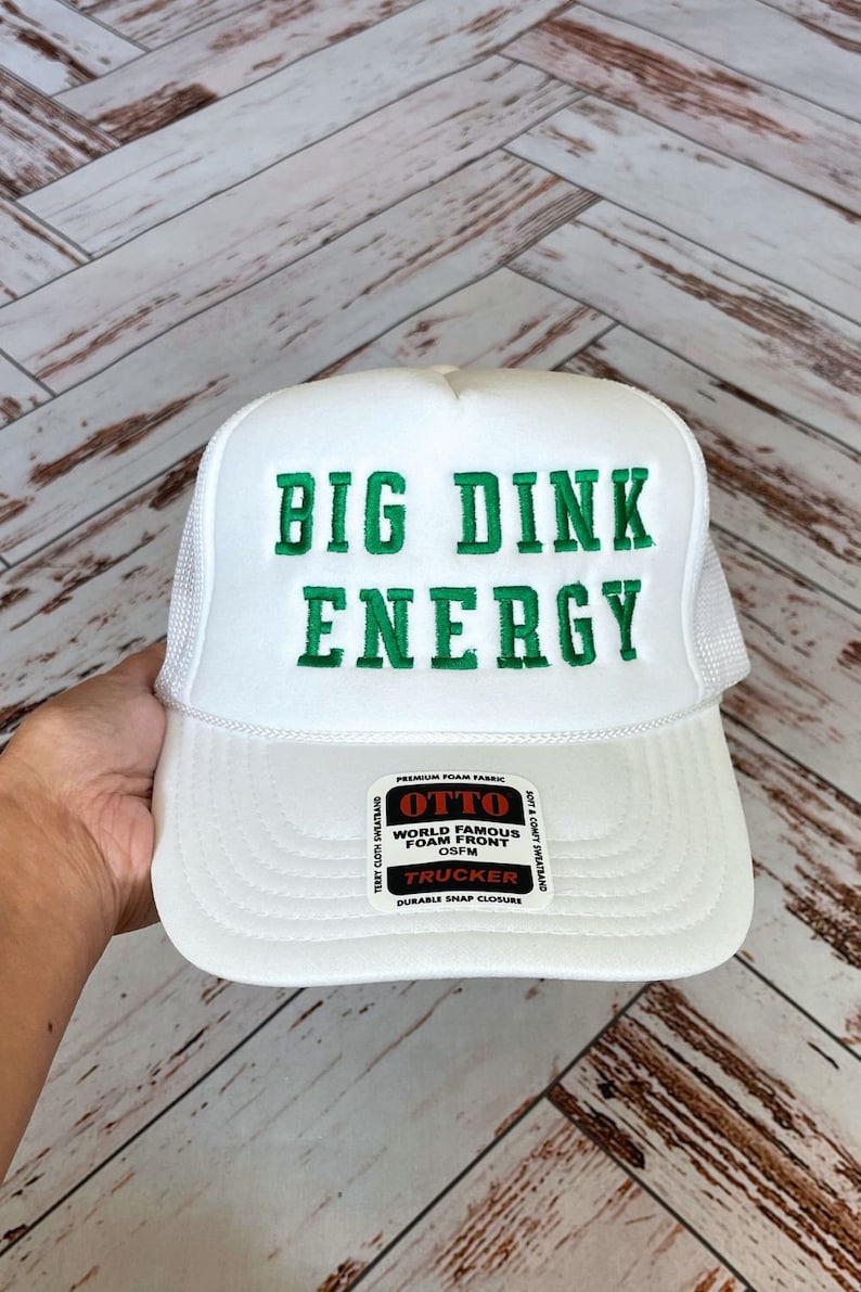 以下が含まれることがあります： 緑色の刺繍で「BIG DINK ENERGY」と書かれた白いトラッカーハット。帽子には、白い文字で「OTTO WORLD FAMOUS FOAM FRONT OSFM TRUCKER DURABLE SNAP CLOSURE」と書かれた黒いラベルが付いています。