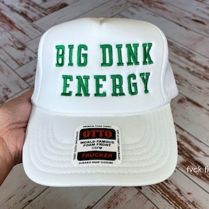 以下が含まれることがあります： 緑色の刺繍で「BIG DINK ENERGY」と書かれた白いトラッカーハット。メッシュバックとスナップクロージャーが付いています。