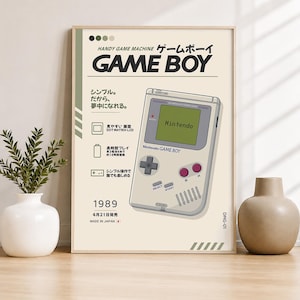 Peut inclure: Une affiche verticale sur fond beige clair présentant une illustration détaillée d'une console de jeu portable grise. Le texte inclut "HANDY GAME MACHINE", "GAME BOY", "1989", "MADE IN JAPAN" et divers caractères japonais.