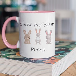 May include: White ceramic mug with a pink rim and handle. The mug has a graphic of three bunnies and the text "Show me your Bunns".