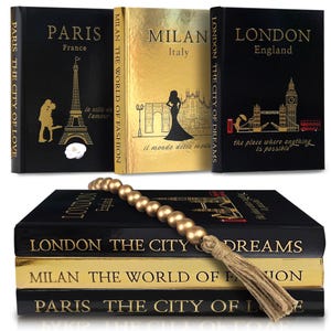 May include: Decorative books with gold lettering and illustrations. The books are black and gold, with titles like "Paris," "Milan," and "London." Each book features city-specific imagery, such as the Eiffel Tower and Big Ben. A gold bead garland with a tassel rests on the books.