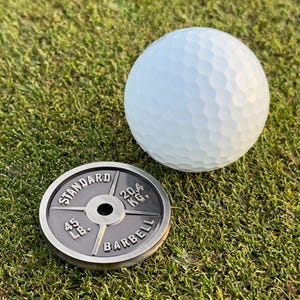May include: A white golf ball with a dimpled surface rests on green grass next to a black and silver barbell weight. The weight is labeled "STANDARD BARBELL" and indicates a weight of 45 lbs and 20.4 kg.