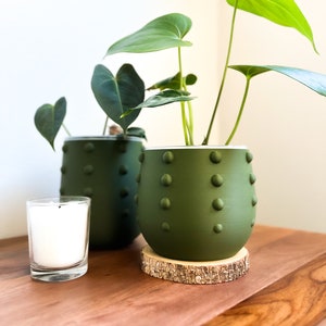 May include: Two green ceramic pots with a textured surface, one with a plant and the other empty, sitting on a wooden surface with a small white candle.