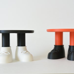 Puede incluir: Dos mesas pequeñas con tapas redondas y patas con forma de pies de dibujos animados. Una mesa tiene una tapa negra y pies blancos, la otra tiene una tapa naranja y pies negros.