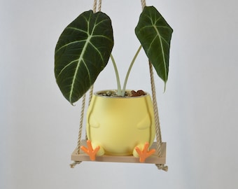May include: A yellow ceramic planter shaped like a chick with orange feet, hanging from a rope swing. The planter holds a green velvet leaf plant.