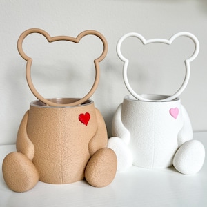May include: Two ceramic bear-shaped planters, one brown and one white, with a heart detail on the front. The planters are sitting on a white surface.