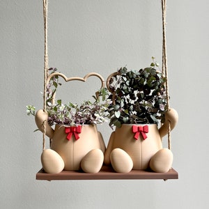 May include: Two beige ceramic bear planters with red bows hanging from a rope swing. Each planter is filled with green and purple trailing plants.