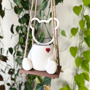 May include: White ceramic planter shaped like a bear with a red heart on its chest, hanging from a brown rope swing.