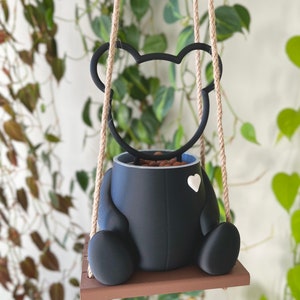 May include: Black bear-shaped hanging planter with a white heart detail. The planter is filled with brown soil and is suspended from a natural rope.