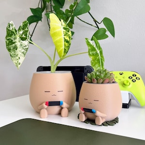 May include: Two beige ceramic planters, each shaped like a character with a face, holding a game controller. One holds a plant with variegated leaves, the other a succulent. A neon green game controller and a black game console are in the background.