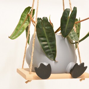 Puede incluir: Una maceta de cerámica gris con forma de pájaro con patas negras, colgada de un columpio de madera con cuerda de yute natural. La maceta está llena de una planta verde de hojas.
