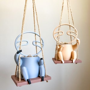 May include: Two hanging planters shaped like sloths, one is light blue and the other is light brown. Both planters have a rope hanging system and a wooden base.