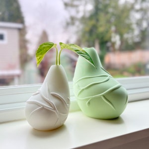 May include: Two pear-shaped ceramic vases with a vine design. The vase on the left is white and the vase on the right is green. A small plant is in the white vase.