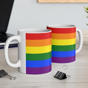 Puede incluir: Dos tazas de cerámica blanca con un diseño de bandera arcoíris. Las tazas tienen un asa blanca y están sobre una superficie de madera clara.