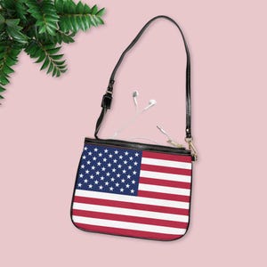 Puede incluir: Un bolso bandolera de cuero negro con cierre de cremallera. El bolso está decorado con un diseño de bandera estadounidense impreso.