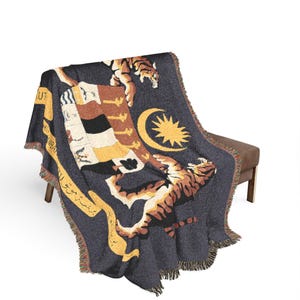 May include: A dark gray woven throw blanket featuring a large, detailed graphic of the Malaysian coat of arms. The design includes two tigers standing on their hind legs, a yellow crescent moon and star, and text in Jawi script along the side.