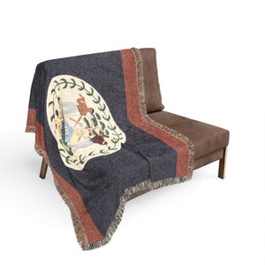May include: A woven throw blanket featuring the Belize national flag design. The blanket has a dark blue centre, red side stripes, and a central white emblem depicting two figures, tools, and a tree. It features a decorative fringed edge.