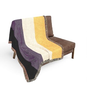 Può includere: Una coperta tessuta con quattro strisce verticali nei colori nero, viola, bianco e giallo. La coperta ha un aspetto lavorato a maglia con bordi sfrangiati multicolori sui lati, drappeggiata sopra una sedia imbottita di colore marrone.