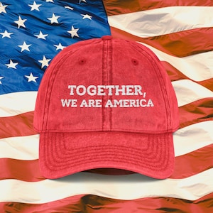 Może przedstawiać: Czerwona czapka bejsbolowa z białym napisem "TOGETHER, WE ARE AMERICA". Czapka jest umieszczona na tle amerykańskiej flagi.