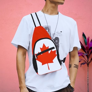 Könnte beinhalten: Eine weiße und rote Umhängetasche mit einem schwarzen Riemen und einem großen roten Ahornblatt-Design. Die Tasche wird von einer Person getragen, die ein weißes T-Shirt trägt.