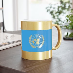 Op de afbeelding: Een gouden koffiemok met een blauw en gouden ontwerp van de vlag van de Verenigde Naties. De vlag toont een wereldkaart omringd door olijftakken.