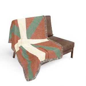 May include: A geometric patterned throw blanket draped over a brown chair. The blanket features a design of alternating stripes in rust, cream, and sage green. The edges are finished with a fringe.