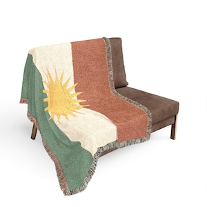 Könnte beinhalten: Eine gewebte Wurfadecke, die über einem braunen Sofa drapiert ist. Die Decke hat ein Design mit grünen, weißen und rostfarbenen Streifen und einem gelben Sonnenstrahl-Design. Die Decke hat einen Fransenrand.