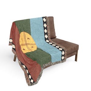 May include: A woven throw blanket featuring a vertical color-blocked design in rust red, dark green, and light blue. A large yellow circular emblem with black line details is centered on the green section. The edges feature a black border with white geometric cross patterns and a fringed hem.