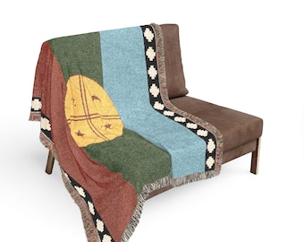 May include: A woven throw blanket featuring a vertical color-blocked design in rust red, dark green, and light blue. A large yellow circular emblem with black line details is centered on the green section. The edges feature a black border with white geometric cross patterns and a fringed hem.