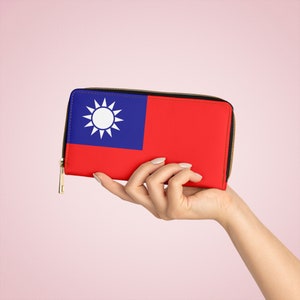 Puede incluir: Un monedero rojo y azul con la bandera de Taiwán. El monedero tiene una cremallera dorada y un emblema de sol blanco. El monedero rectangular es sostenido por una mano.