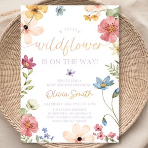 Puede incluir: Una invitación a una baby shower con un diseño floral de acuarela. El texto dice "¡Una pequeña flor silvestre está en camino!" Únase a nosotros para una baby shower en honor a Olivia Smith. Sábado 17 de agosto a las 2:00 PM. La Residencia Hudson, 25 Montreal Ave, Regina, CA. RSVP a Emma al 900-123-4567. Registrado en Target.