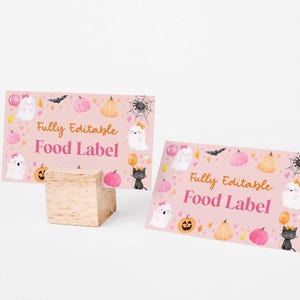 May include: Two pink Halloween-themed food label cards with illustrations of ghosts, pumpkins, bats, and spiders. The cards have the text "Fully Editable Food Label" in orange and pink. One card is propped up on a wooden block.