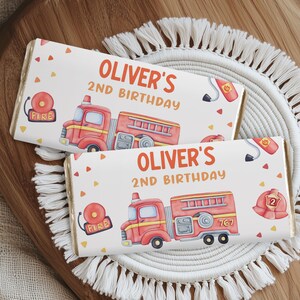 Puede incluir: Dos envoltorios de barra de chocolate personalizados con un tema de camión de bomberos. Los envoltorios son blancos con acentos rojos y amarillos. El texto en los envoltorios dice "Oliver's 2nd Birthday".