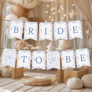 May include: A decorative banner with the words "BRIDE TO BE" in blue letters. The banner is strung across a room decorated for a bridal shower. The background includes curtains, balloons, and decorative items.