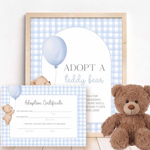 May include: A brown teddy bear sits next to a framed print and an adoption certificate. The print features the text "ADOPT A teddy bear" with a blue balloon graphic. The certificate has space for names and promises to love and care for a new bear.