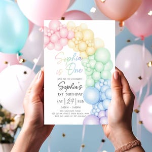 May include: A pastel-colored birthday invitation held by hands, featuring a rainbow balloon arch design. The invitation reads "Sophia is One" and includes event details: date, time, and location. Balloons in various pastel shades fill the background.