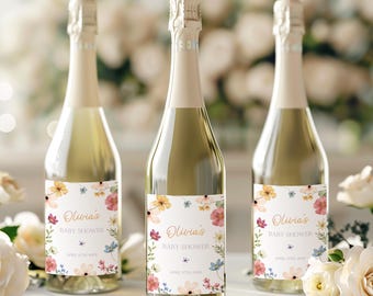 Könnte beinhalten: Drei Champagnerflaschen mit personalisierten Etiketten für eine Babyparty. Die Etiketten zeigen ein Aquarell-Blumendesign mit rosa, gelben und blauen Blumen. Der Text auf den Etiketten lautet "Olivia's Baby Shower" mit dem Datum "April 17th, 2023".