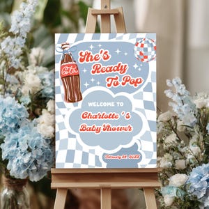 May include: A baby shower sign on a wooden easel. The sign features a retro design with the text "She's Ready To Pop" and "Welcome to Charlotte's Baby Shower". A vintage Coca-Cola bottle graphic is on the left.