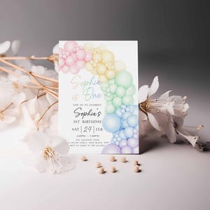 May include: A white invitation card with a pastel rainbow balloon design. The text reads "Sophia is One" and "Sophia's 1st Birthday". The card is decorated with white flowers and small, light-colored beads.
