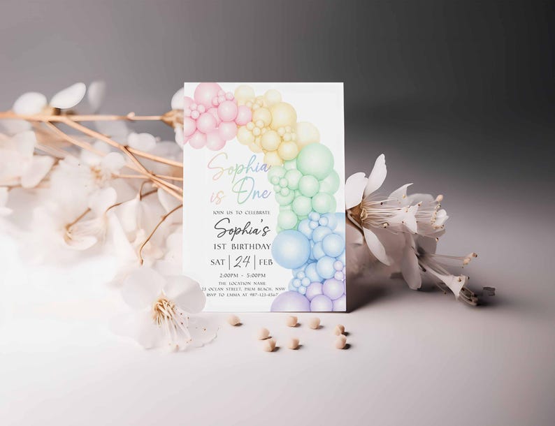 May include: A white invitation card with a pastel rainbow balloon design. The text reads "Sophia is One" and "Sophia's 1st Birthday". The card is decorated with white flowers and small, light-colored beads.