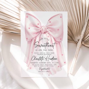 May include: A baby shower invitation featuring a large pink bow adorned with small pink hearts. The text "Our Little Sweetheart Is On The Way!" is displayed, along with the honoree's name, date, time, and RSVP information.