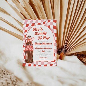 May include: A baby shower invitation with a retro design. The invitation features a Coca-Cola bottle illustration and the text "She's Ready To Pop." The background has a red and pink checkered pattern. The invitation includes the date, time, and location.