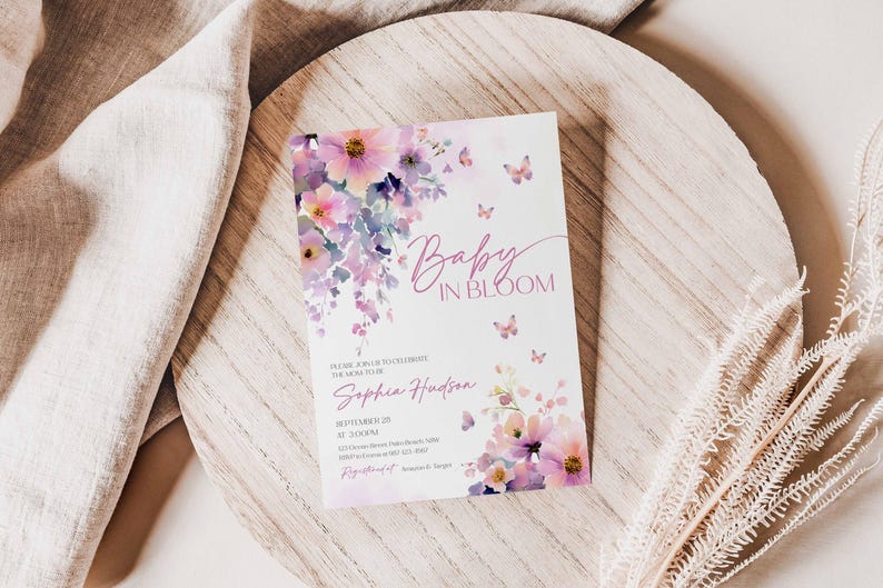 Może przedstawiać: Zaproszenie na baby shower z ilustracjami kwiat&oacute;w i motyli. Zaproszenie zawiera tekst "Baby in Bloom" w czcionce skryptowej, wraz ze szczeg&oacute;łami wydarzenia. Projekt zawiera odcienie r&oacute;żu, fioletu i bieli.